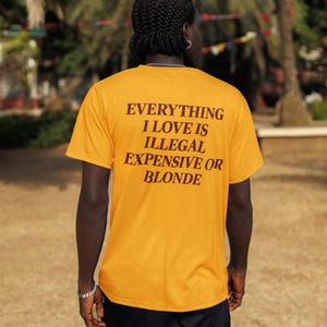 May include: A mustard yellow short-sleeve t-shirt featuring dark brown text on the back that reads: EVERYTHING I LOVE IS ILLEGAL EXPENSIVE OR BLONDE. The shirt is paired with black cargo shorts, creating a casual look with a bold graphic statement.