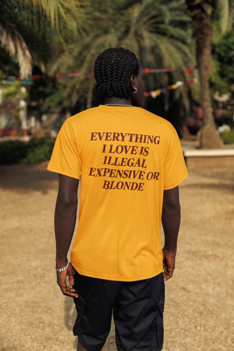 May include: A mustard yellow short-sleeve t-shirt featuring dark brown text on the back that reads: EVERYTHING I LOVE IS ILLEGAL EXPENSIVE OR BLONDE. The shirt is paired with black cargo shorts, creating a casual look with a bold graphic statement.