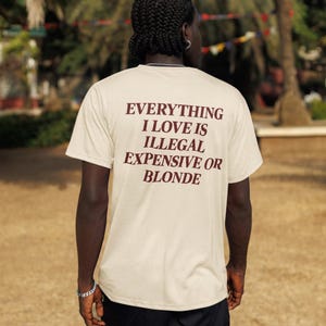 May include: A beige short-sleeved t-shirt featuring dark brown text on the back that reads: EVERYTHING I LOVE IS ILLEGAL EXPENSIVE OR BLONDE. The shirt has a crew neckline and a standard fit, shown from the back view in an outdoor setting.