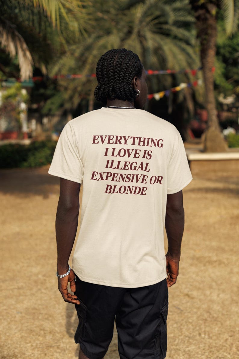 May include: A beige short-sleeved t-shirt featuring dark brown text on the back that reads: EVERYTHING I LOVE IS ILLEGAL EXPENSIVE OR BLONDE. The shirt has a crew neckline and a standard fit, shown from the back view in an outdoor setting.