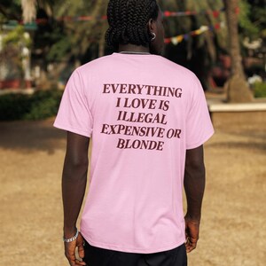 May include: A light pink short-sleeved t-shirt featuring dark brown text on the back that reads: EVERYTHING I LOVE IS ILLEGAL EXPENSIVE OR BLONDE. The shirt is paired with black cargo shorts, creating a casual look with clear, bold lettering.