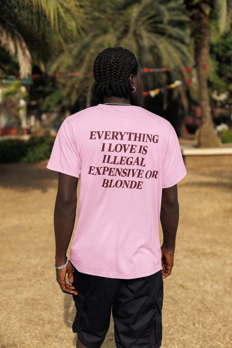 May include: A light pink short-sleeved t-shirt featuring dark brown text on the back that reads: EVERYTHING I LOVE IS ILLEGAL EXPENSIVE OR BLONDE. The shirt is paired with black cargo shorts, creating a casual look with clear, bold lettering.