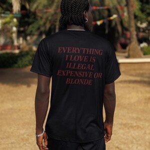 May include: A black short-sleeved t-shirt featuring the text "EVERYTHING I LOVE IS ILLEGAL EXPENSIVE OR BLONDE" printed in dark red lettering on the back. The shirt is paired with black cargo shorts, creating a simple and functional outfit.