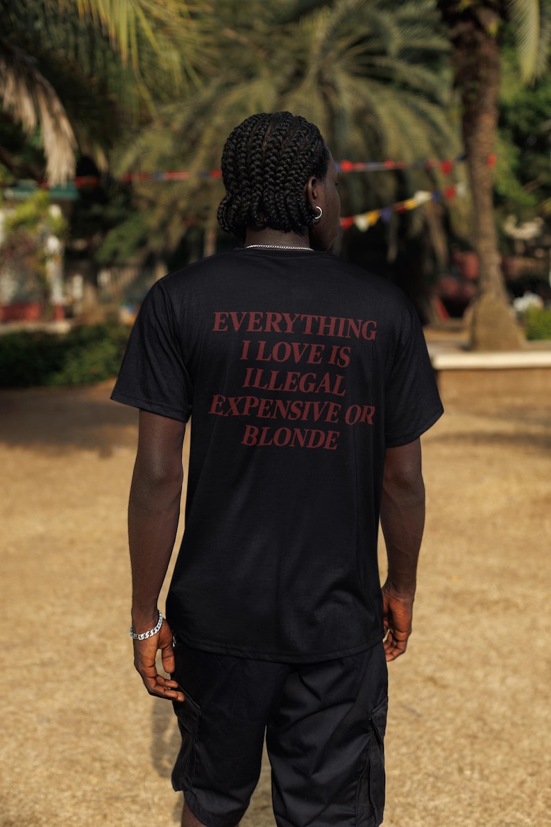 May include: A black short-sleeved t-shirt featuring the text "EVERYTHING I LOVE IS ILLEGAL EXPENSIVE OR BLONDE" printed in dark red lettering on the back. The shirt is paired with black cargo shorts, creating a simple and functional outfit.