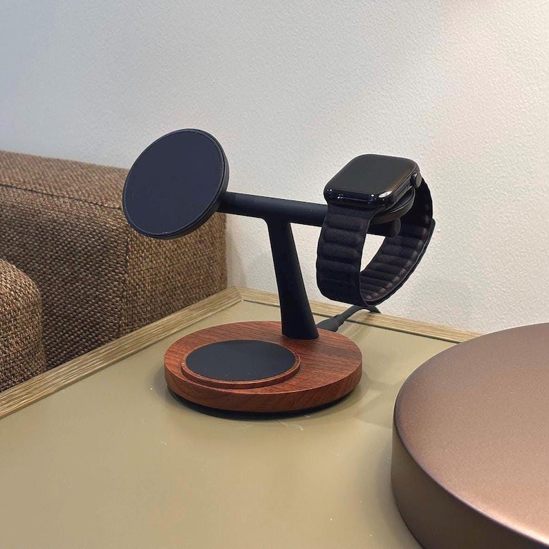 Op de afbeelding: Een houten oplaadstation met een zwarte T-vormige arm die een zwarte smartwatch vasthoudt. De basis is cirkelvormig met een donkere houten afwerking en een zwart oplaadpad. Een zwart cirkelvormig oplaadpad bevindt zich aan de linkerkant.