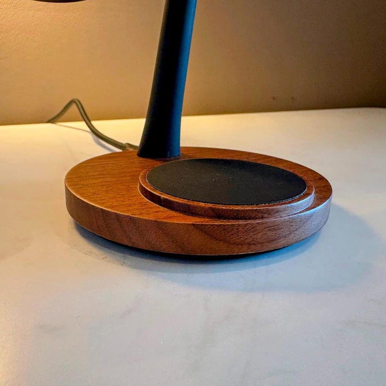 May include: A modern desk lamp with a wooden base and a black stem. The base is a rich brown color with a circular design, and a black pad is set in the center. The lamp's design is minimalist and stylish.