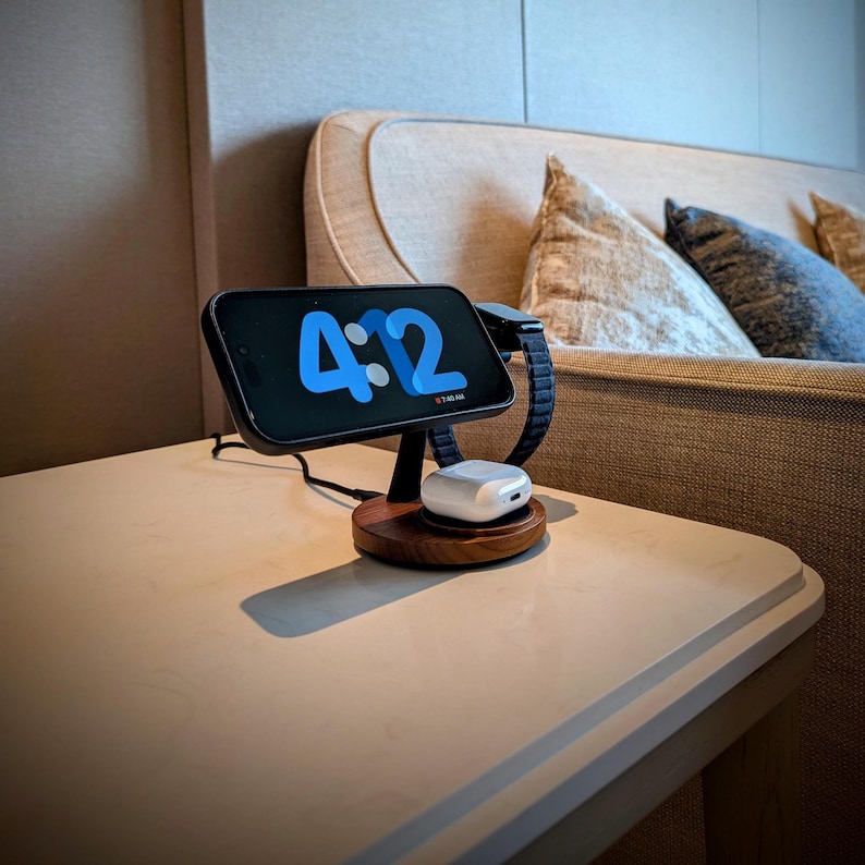 May include: A wooden charging station with a black smartphone displaying the time 4:2, a black smartwatch, and white earbuds. The charging station is on a light-colored table, with a sofa in the background.