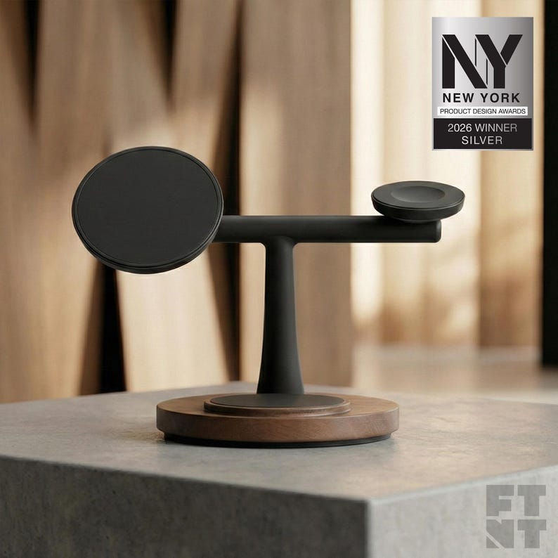 Op de afbeelding: Een zwart en houtkleurig oplaadstation met een cirkelvormige basis en een horizontale arm met twee cirkelvormige oplaadpads. Het ontwerp is modern en minimalistisch. De afbeelding bevat een zilveren award-badge met de tekst "NY New York Product Design Awards 2026 Winner Silver."