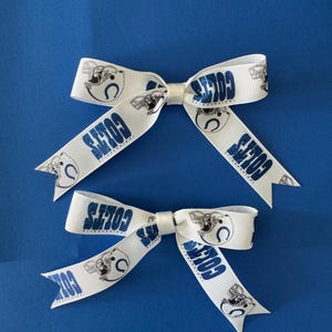 May include: Two white hair bows made of ribbed fabric featuring a repeating blue and black print of the Indianapolis Colts football team logo, including the horseshoe emblem and the word Colts in a bold blue font on the ribbon tails.