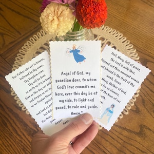 Può includere: Tre biglietti di preghiera con bordi smerlati sono tenuti davanti a un vaso di fiori. Le carte presentano testo religioso e illustrazioni di un angelo e della Vergine Maria. I fiori includono fiori arancioni, gialli e rosa.