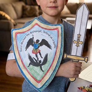 Puede incluir: Un conjunto de escudo y espada de papel con una ilustración coloreada a mano del Arcángel San Miguel. El escudo muestra el texto Saint Michael the Archangel, Defend us in battle, con detalles en rojo, azul y dorado. La espada tiene una cruz dorada.