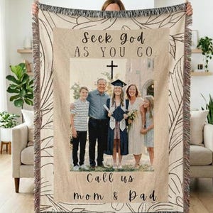 Könnte beinhalten: Eine gewebte Wohndecke mit einem zentralen Foto einer vierköpfigen Familie, inklusive einer Person im Abschlussgewand. Der Text Seek God AS YOU GO und Call us mom & Dad ist schwarz aufgedruckt. Der Rand hat ein beiges Blattmuster und Fransen.