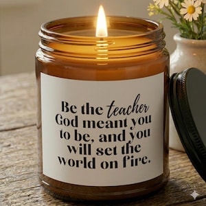 Può includere: Una candela accesa in un barattolo di vetro marrone con coperchio nero. La candela ha un'etichetta bianca con il testo "Be the teacher God meant you to be, and you will set the world on fire."