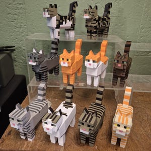 May include: A collection of ten blocky, pixelated cat figurines in various colors and patterns, including grey tabby, orange tabby, white, and brown striped designs. Each cat features a square head, pointed ears, and a vertical tail.
