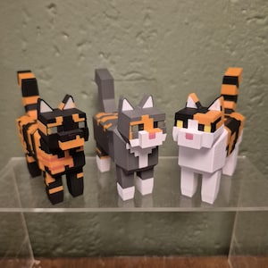 Puede incluir: Tres figuras de gato cúbicas y pixeladas colocadas una al lado de la otra. El gato de la izquierda es negro y naranja, el del medio es gris, blanco y naranja, y el de la derecha es blanco, negro y naranja. Cada gato tiene cabeza y cola cuadradas.