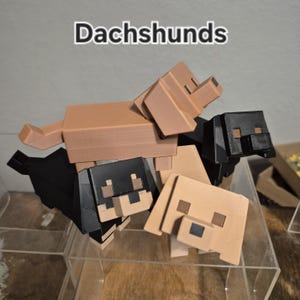 May include: A collection of four blocky, pixelated dachshund figurines in tan and black colors. The figures feature square heads, rectangular bodies, and short legs. The word Dachshunds is printed in white text with a black outline above the dogs.