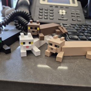 May include: Four blocky, pixelated cat figurines in black, white, brown, and tan. Each cat features a square head with distinct colored eyes, a small nose, and a rectangular body with short legs. The figures are arranged in a group on a flat surface.