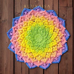 May include: A colorful, handmade crochet mandala in a rainbow of hues, including green, yellow, pink, and blue. The intricate design features a central floral pattern and a scalloped edge. The word "KUUKAISSA" is visible.