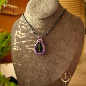 Op de afbeelding: Een handgemaakte ketting met een donkergroene hanger, omlijst door paars en geel macramé, hangt aan een grijze display buste. De ketting heeft een gevlochten zwart koord. De merknaam "KUUKAIS" is onderaan zichtbaar.
