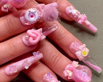 May include: A set of long, pink acrylic nails with various 3D embellishments. Designs include a pink orb filled with glitter, a pink eye with pearl accents, a pink pom-pom, and a pink teddy bear. Other nails feature bows, flowers, and gems.