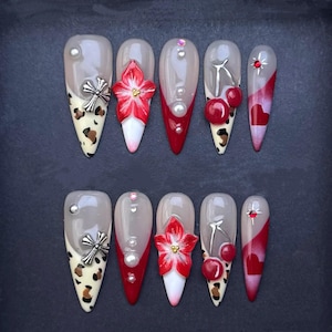 May include: A set of ten long, almond-shaped artificial nails. Designs include a mix of leopard print, red accents, silver embellishments, and 3D floral and cherry details. Some nails feature pearls and a cross design.