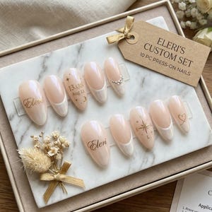 May include: A set of ten Eleri's Custom Press-On Nails in a display box. The nails are a light pink with white tips, some adorned with gold text, a heart, a starburst, and rhinestones. A tag reads "Eleri's Custom Set 10 PC Press-On Nails."