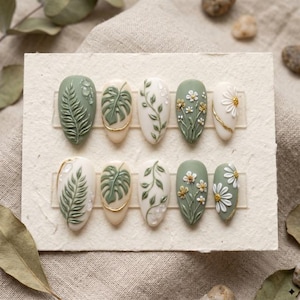 Puede incluir: Un conjunto de diez uñas artificiales con diseños botánicos. Las uñas presentan patrones de hojas de helecho, hojas de monstera y arreglos florales. La paleta de colores incluye tonos verdes, blancos y acentos dorados.