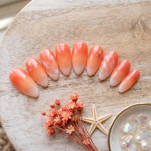 Puede incluir: Un juego de diez uñas postizas en forma de almendra con un diseño degradado de naranja a blanco, que incluye sutiles remolinos tipo mármol blanco y pequeñas escamas de purpurina iridiscente incrustadas en el acabado brillante de cada uña.
