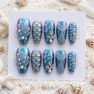 May include: A set of ten iridescent press-on nails with a mermaid theme. Designs include shell, scale, bubble, coral, mermaid tail, and wave patterns. Embellishments include pearls and small crystals. The nails are displayed on a white card.