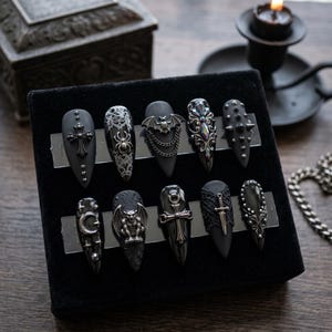 May include: A set of ten black press-on nails with pointed tips, featuring silver-toned embellishments including a cross, spider, bat with chains, ornate filigree, studs, crescent moon, gargoyle, sword, and intricate lace-like patterns on a display.