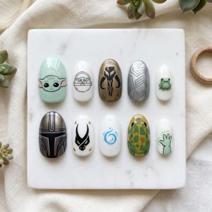 May include: A collection of ten artificial nails, each decorated with a different design. The designs feature a character with large eyes, a character in a pod, a symbol, a geometric pattern, a frog, a helmet, a symbol, a swirl, a turtle, and a hand.