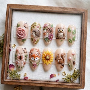 May include: A set of ten almond-shaped nail art designs. Each nail showcases a unique 3D design, such as a rose, butterfly, hedgehog, flowers, owl, leaves, fox, sunflower, squirrel, and ladybug. The nails are presented in a wooden frame.