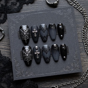 May include: A set of ten press-on nails in a coffin shape, featuring a black base with silver spider web patterns, silver cross charms, intricate black lace designs, small silver skull accents, and polished black surfaces with silver studs.