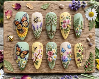 May include: A collection of ten artificial nails with intricate, 3D floral and insect designs. Each nail features a different design, including butterflies, bees, flowers, and trees, in a variety of colors. The nails are arranged on a wooden surface.