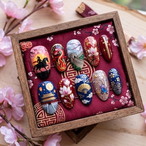 May include: A set of ten press-on nails displayed in a wooden box on a red fabric cushion. Each nail features detailed hand-painted designs including a warrior on horseback, a red dragon, the Great Wall, lotus flowers, a sword, a palace, a fan, and a cricket.