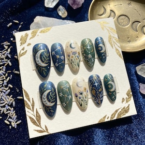 May include: A set of ten press-on nails with celestial designs. Each nail features a different design, including moons, stars, and floral patterns, in shades of blue, green, and cream. The nails are displayed on a white card with gold leaf accents.