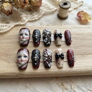 May include: A set of ten press-on nails featuring intricate designs including a doll face with cracked porcelain texture, black lace patterns with white pearls, silver charms, black bows, and deep red nails with silver stitch-like accents.