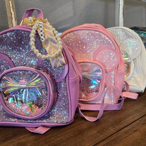 May include: Four sparkly mini backpacks with clear plastic front pockets and a bow design. The backpacks are pink, purple, white, and black. The purple backpack has a name tag that says "Jenna".