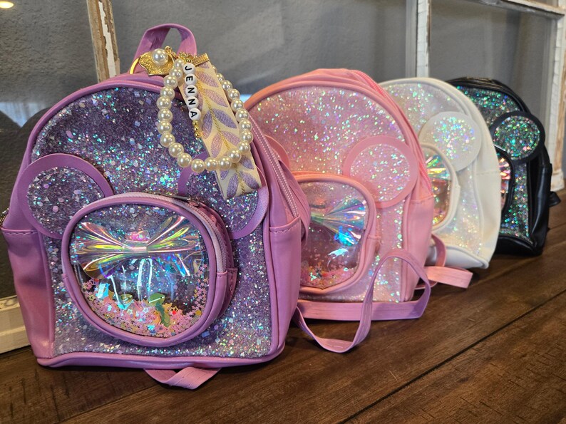 May include: Four sparkly mini backpacks with clear plastic front pockets and a bow design. The backpacks are pink, purple, white, and black. The purple backpack has a name tag that says "Jenna".