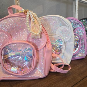 May include: Four sparkly mini backpacks in pink, white, pink, and black. The pink backpack has a clear plastic window with a rainbow bow and glitter inside. The backpack has a pearl and letter charm that says "Chloe".