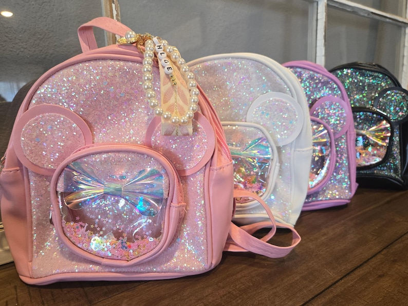 May include: Four sparkly mini backpacks in pink, white, pink, and black. The pink backpack has a clear plastic window with a rainbow bow and glitter inside. The backpack has a pearl and letter charm that says "Chloe".