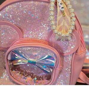 May include: Pink glitter backpack with a clear pocket featuring a rainbow bow and glitter. The backpack has a pearl and ribbon charm with the word "LOVE" on it.