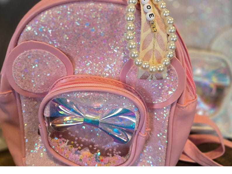 May include: Pink glitter backpack with a clear pocket featuring a rainbow bow and glitter. The backpack has a pearl and ribbon charm with the word "LOVE" on it.