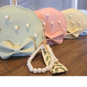 May include: Four pastel-colored faux-shell purses with pearl accents and a bow detail. The purses are attached to a pearl and green leaf charm.