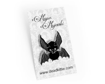 Könnte beinhalten: Schwarzer Fledermaus-Emaille-Pin mit detaillierten Flügeln und einem niedlichen Gesicht, auf einer gemusterten Karte. Die Karte trägt den Namen "Megan Majewski" und die Website "www.deadkitti.com".