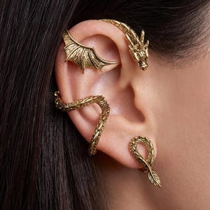 Op de afbeelding: Goudkleurige oorjuwelen met een draakthema. De set bevat een drakenkop, een vleugel en een slangachtig ontwerp dat zich om het oor wikkelt. De sieraden worden op het oor gedragen en zijn zichtbaar op de huid.