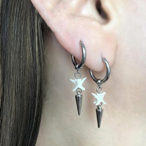 May include: Silver hoop earrings with white bone-shaped charms and dark gray cone pendants. The earrings are shown on an ear, with the focus on the jewelry's design and details. The earrings are a unique accessory.