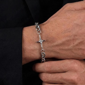 May include: A silver bracelet with a cross-shaped charm featuring a central gemstone. The bracelet is made of linked chain segments and is worn on a wrist. The background is a dark suit.