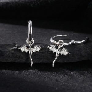 May include: A pair of silver hoop earrings with a dragon charm. Each earring features a small, detailed dragon pendant with outstretched wings suspended from a circular hoop. The earrings are displayed against a dark, textured background.