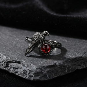 Peut inclure: Bague en argent foncé ornée d'un corbeau perché au-dessus d'une pierre rouge ronde. La bague présente des détails complexes sur l'oiseau et le sertissage, créant une esthétique gothique ou fantastique. La bague est présentée sur une surface texturée gris foncé.